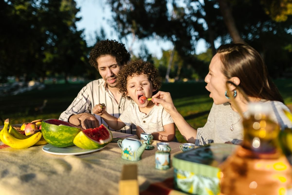 Gezin eet samen buiten in een zonnig groen park, sfeerbeeld bij de restaurants en Market Dome van Center Parcs Port Zélande