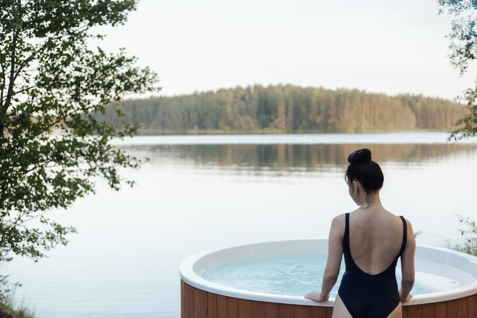 Vrouw stapt in een hottub aan een meer met bos, vakantiehuisje met hottub